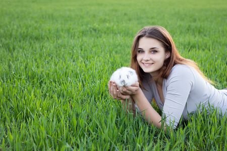 Girl holding a rabbit while on a green meadow. Young smiling girl holding a little fluffy white rabbit. Friendship with Easter Bunny. Spring photo with beautiful young girl with her Bunnyの写真素材