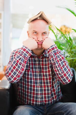 A bald European man in a crippled red shirt and blue classic jeans is sitting in an armchair in a bright apartment with green plants, holding a book over his head. Tired of studying manの写真素材