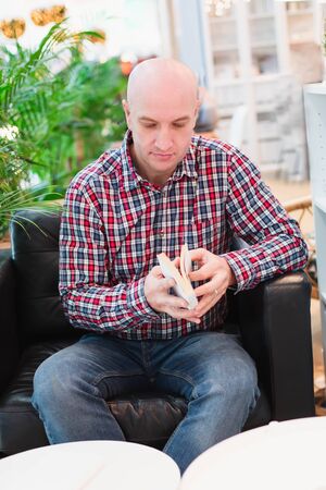 A bald European man in a checkered red shirt and blue jeans sits in an armchair in a bright apartment with green plants, chooses books to read in his free time. Man engage in self-developmentの写真素材