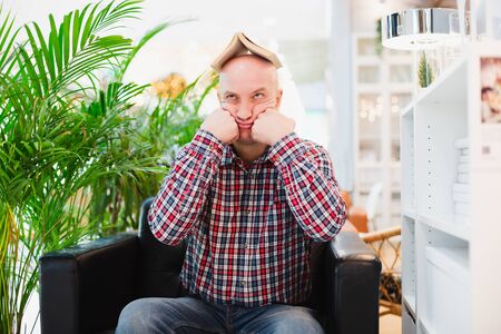 A bald European man in a crippled red shirt and blue classic jeans is sitting in an armchair in a bright apartment with green plants, holding a book over his head. Tired of studying manの写真素材