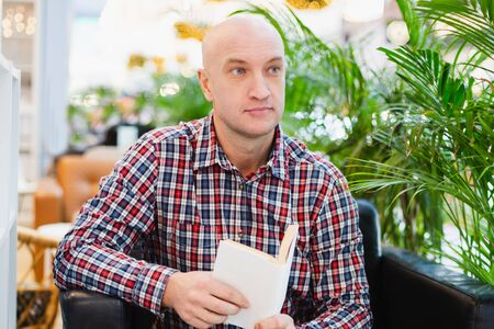 bald european man in a red checkered shirt and blue jeans sits in an armchair in a bright apartment with a lot of green plants, reads a book in a home relaxing interiorの写真素材