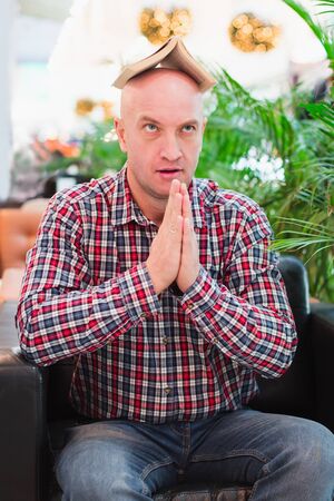 A bald European man in a crippled red shirt and blue classic jeans is sitting in an armchair in a bright apartment with green plants, holding a book over his head. Tired of studying manの写真素材