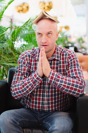 A bald European man in a crippled red shirt and blue classic jeans is sitting in an armchair in a bright apartment with green plants, holding a book over his head. Tired of studying manの写真素材