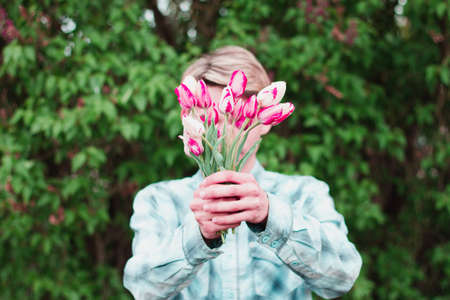 guy in a blue checkered shirt holds a bouquet of pink tulips holding them in front of him, close his face, outdoors.の写真素材
