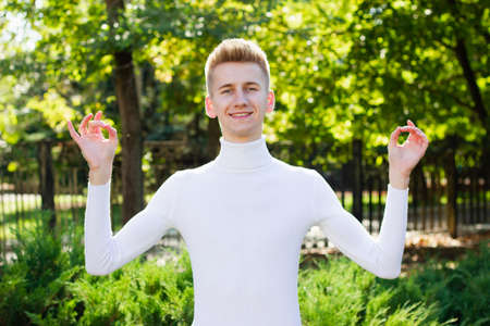 Portrait of a cheerful young man, dressed in white clothes, and showing ok gesture with his hands, isolated from everyone in a city park.の写真素材