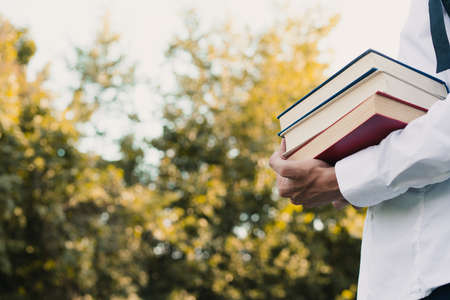 a guy without a face holds a stack of books in one hand against the background of a green park, the concept of new knowledge. Place for textの写真素材