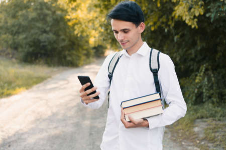 a young european guy in a white classic shirt with a gray backpack on his back holds a stack of books in his hand and looks into his phone, outdoors. education conceptの写真素材