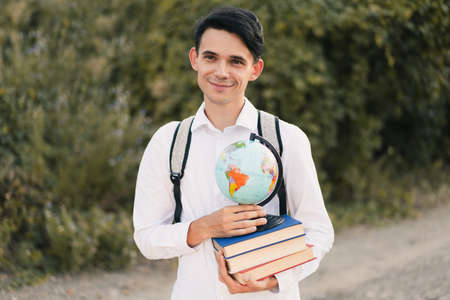 young European guy in a white classic shirt with a gray backpack on his back holds a stack of educational books with globe and smiles cutely against the background of green nature. Education conceptの写真素材