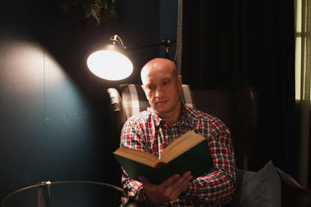 A man, dressed in a plaid shirt and jeans, sits in an armchair and reads a book at dusk. Artificial lighting. Life concept.の写真素材