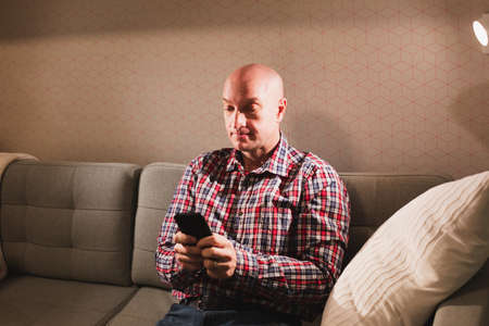 Portrait of a happy adult bald man, dressed in a plaid shirt and jeans, sitting on the couch with a phone.の写真素材