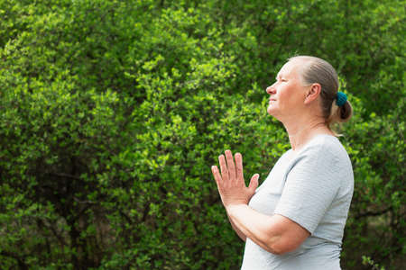 A calm elderly woman, dressed in white clothes, meditates in nature. Practice yoga, relieve stress or renew energy. Healthy lifestyle concept.の写真素材