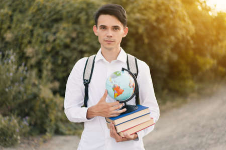 young European guy in a white classic shirt with a gray backpack on his back holds a stack of educational books with globe and smiles cutely against the background of green nature. Education conceptの写真素材