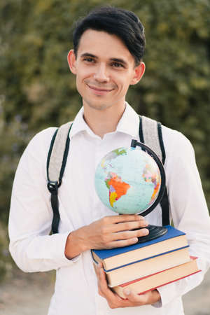 young European guy in a white classic shirt with a gray backpack on his back holds a stack of educational books with globe and smiles cutely against the background of green nature. Education conceptの写真素材
