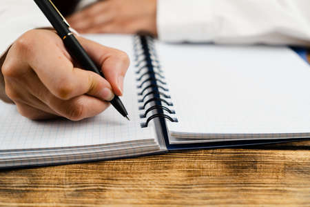 A schoolgirl, dressed in a white blouse, sits at a wooden desk and holds in her hand a black pen over a notebook. School concept.の写真素材