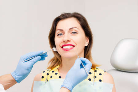 The young face of a smiling woman, wearing a yellow blouse and bright lipstick, is being worked on by a dentist wearing blue gloves. Close-up.の写真素材