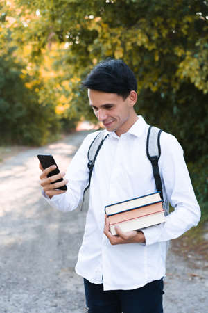 A dark-haired schoolboy, dressed in a white and clean shirt, with a gray briefcase on his shoulders, holds a phone and multi-colored books on an autumn background.の写真素材