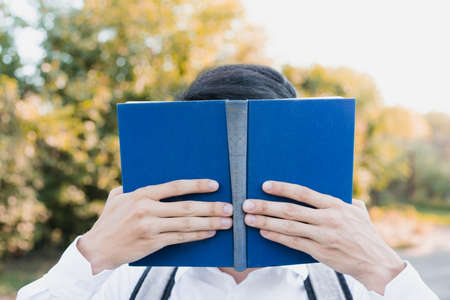 A dark-haired schoolboy, dressed in a white shirt, with a briefcase on his shoulders, holds a large blue book with both hands and covers his face against an autumn background.の写真素材