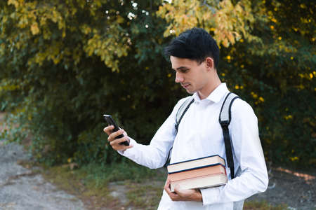 A dark-haired schoolboy, dressed in a white and clean shirt, with a gray briefcase on his shoulders, holds a phone and multi-colored books on an autumn background.の写真素材