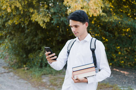 A dark-haired schoolboy, dressed in a white and clean shirt, with a gray briefcase on his shoulders, holds a phone and multi-colored books on an autumn background.の写真素材