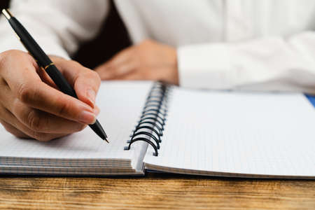 A schoolgirl, dressed in a white blouse, sits at a wooden desk and holds in her hand a black pen over a notebook. School concept.の写真素材
