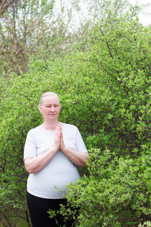 A mature woman, dressed in white clothes, holding hands, resting after yoga alone in the morning. Concentrated on relaxation. Healthy life concept.の写真素材