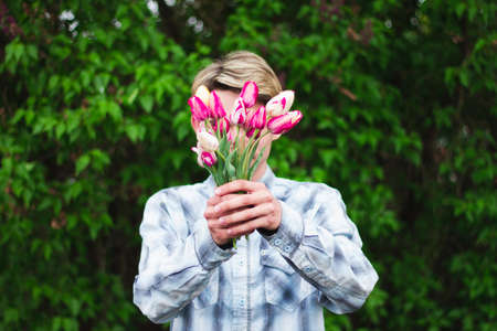 guy in a blue checkered shirt holds a bouquet of pink tulips holding them in front of him, close his face, outdoors.の写真素材