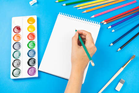 A schoolboy holds a pencil over a notebook on a blue background and school supplies. School concept. View from above.の写真素材