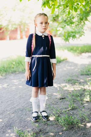 Little schoolgirl girl, dressed in a blue uniform, with two pigtails and a pink backpack on her shoulders, in white socks on an autumn background. School concept.の写真素材