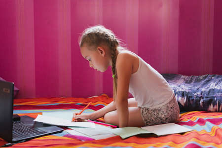 A little girl of Caucasian appearance in home casual clothes does tasks in front of a laptop, drawing on a white sheet with colored pencils. The concept of social isolation during the epidemicの写真素材