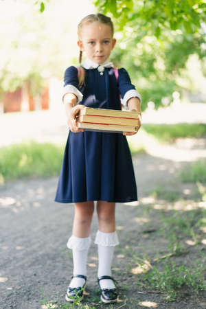 a young girl of European appearance in a dark blue school uniform with a backpack on her back holds a stack of books in front of her. Concept for the first day of schoolの写真素材