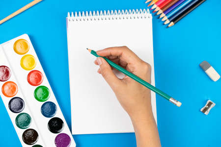A schoolboy holds a pencil over a notebook on a blue background and school supplies. School concept. View from above.の写真素材
