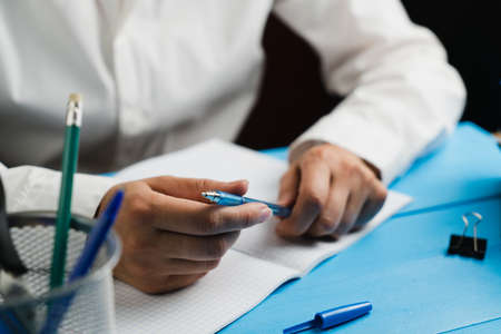 A schoolgirl in a white shirt sits at a wooden table with an empty notebook and holds a blue pen with both hands. School concept. Close-up.の写真素材