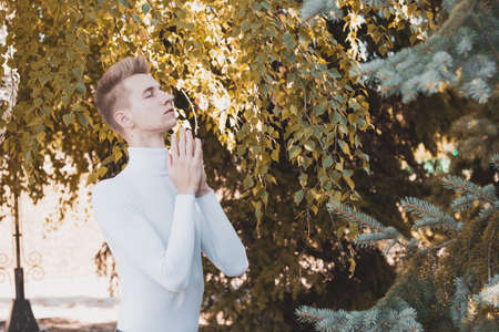 A young European, a young blond man, dressed in a white jacket, holds his hands in front of him, folding their palm to palm, with closed eyes against a warm autumn background.の写真素材