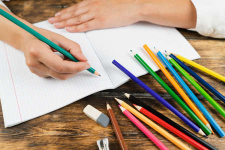 A schoolgirl, dressed in a white blouse, sits at a wooden desk and draws in a squared notebook with colored pencils. School concept.の写真素材