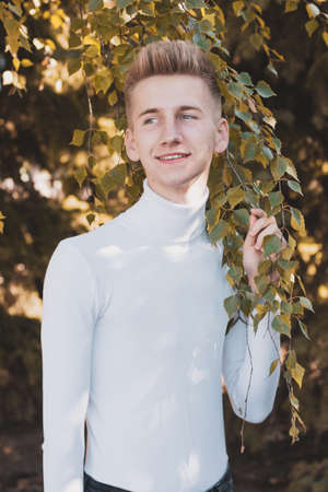 A blue-eyed young blond European guy, dressed in a white sweater and black jeans, stands against a warm autumn background. Life concept.の写真素材