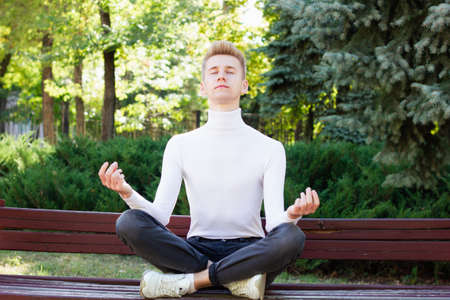 A male athlete, dressed in a white sweater and black jeans, sits in a lotus position, resting and meditating after yoga. Summer in the park on the bench. Healthy life concept.の写真素材
