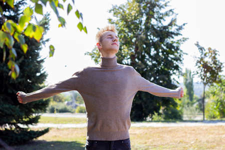 A young blond European guy, dressed in a bodysuit, stands with outstretched hands against the background of autumn nature. Photo concept.の写真素材