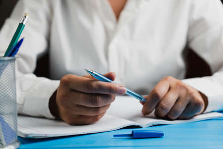A schoolgirl in a white and clean shirt sits at a wooden table with an empty notebook and holds a blue pen with both hands. School concept.の写真素材