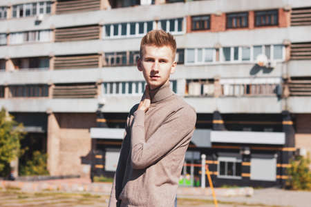 Young European blond guy posing against the background of a multi-storey building on an ordinary autumn day. Photo concept.の写真素材