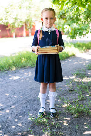 A little schoolgirl girl with two pigtails, dressed in a blue uniform, with a pink backpack on her shoulders and white socks, holds books in her hands against the background of nature.の写真素材