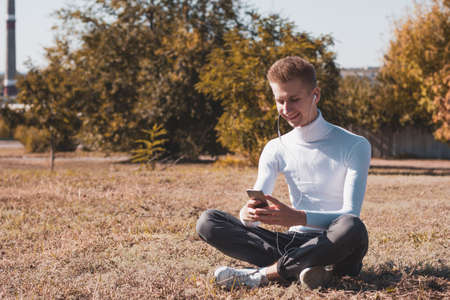 A young blond guy, dressed in a white sweater and black jeans, sits on the ground and listens to music using headphones and a phone against the background of autumn trees.の写真素材