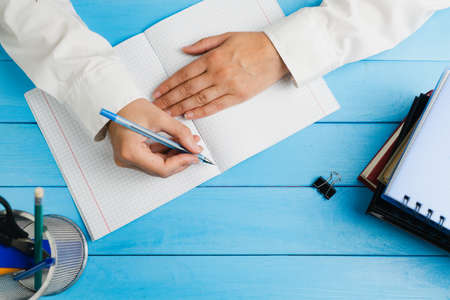 A schoolgirl, dressed in a white dress shirt, sits at a blue wooden table with school supplies and holds a pen over a notebook. View from above.の写真素材