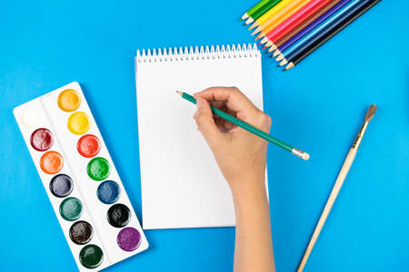 A schoolboy holds a pencil over a notebook on a blue background and school supplies. School concept. View from above.の写真素材