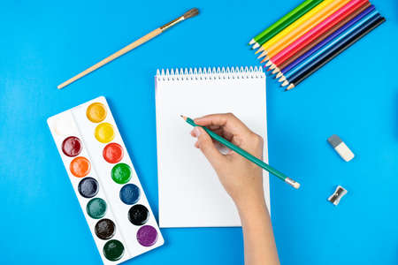 A schoolboy holds a pencil over a notebook on a blue background and school supplies. School concept. View from above.の写真素材