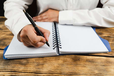 A schoolgirl, dressed in a white blouse, sits at a wooden desk and holds in her hand a black pen over a notebook. School concept.の写真素材