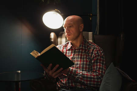 An adult man learns by reading a book in focus at night by the light of a lamp. A dark room.の写真素材