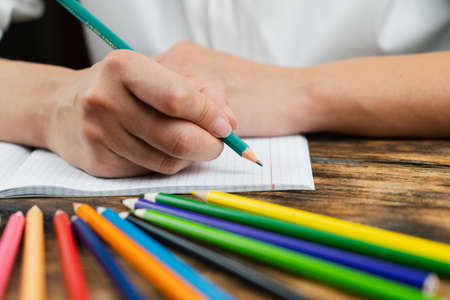 A schoolgirl, dressed in a white blouse, sits at her desk with a notebook against the background of colored pencils. Close-up.の写真素材