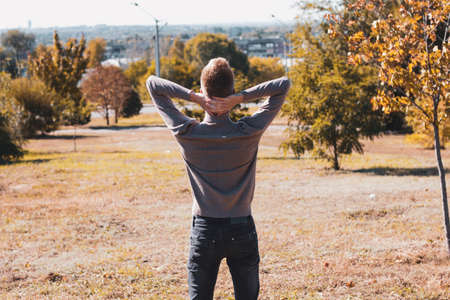 A young blond guy, dressed in a beige sweater and black jeans, stands with his back to the camera and holds his hands behind his head against the background of autumn trees.の写真素材