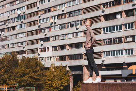 A young blond guy, dressed in a beige sweater and black jeans, stands half-side with closed eyes against the backdrop of a multi-storey building. Photo concept.の写真素材