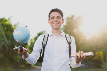 Smiling brunette guy with gray briefcase on his shoulders, dressed in  classic white shirt, stands and holds globe in one hand, and in the second book against the background of nature and sunset.の写真素材